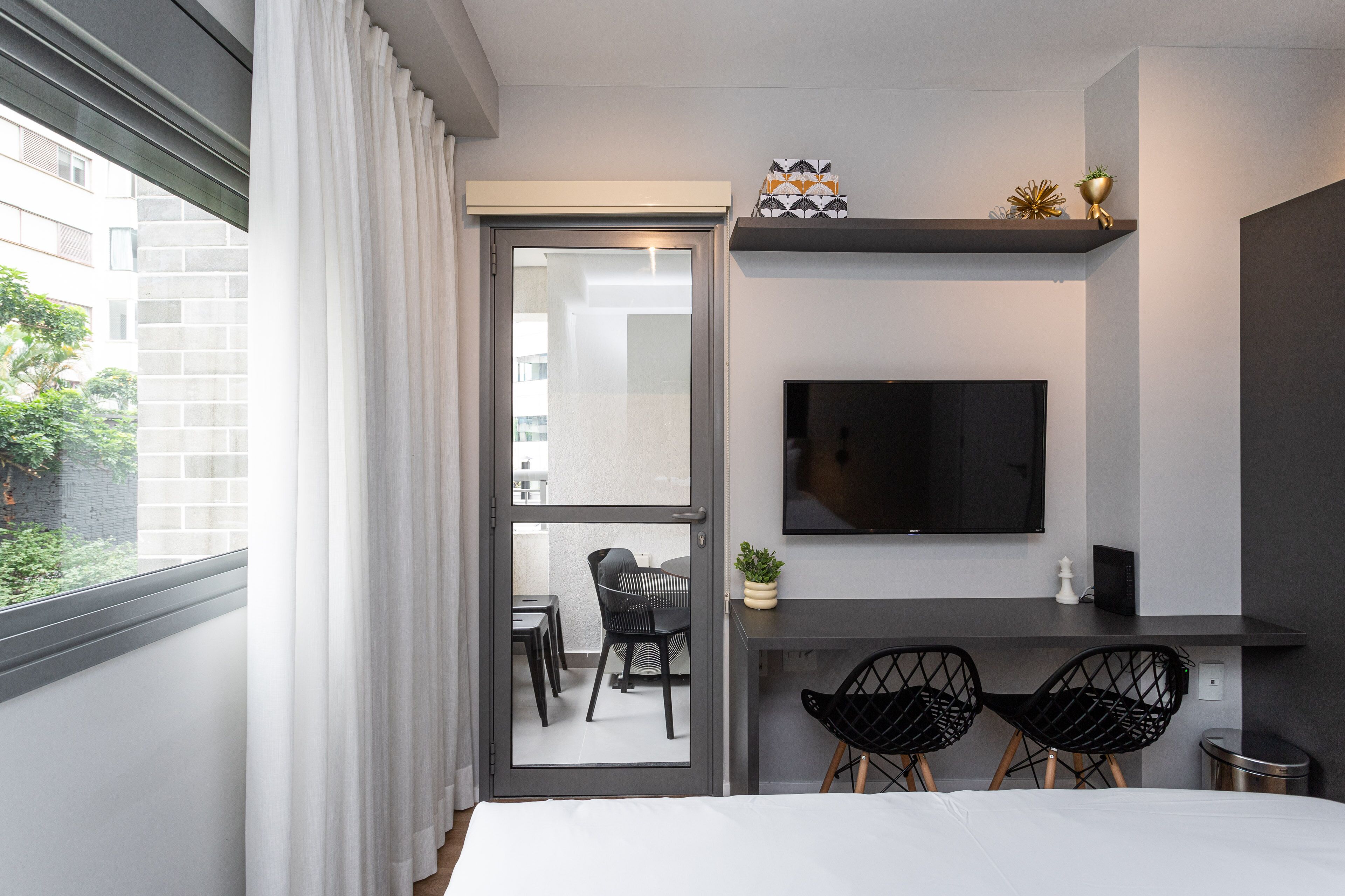 Une chambre d’hôtel moderne équipée d’un lit, d’un bureau avec deux chaises, d’une télévision et d’une porte-fenêtre coulissante donnant sur un balcon avec une table et des chaises.