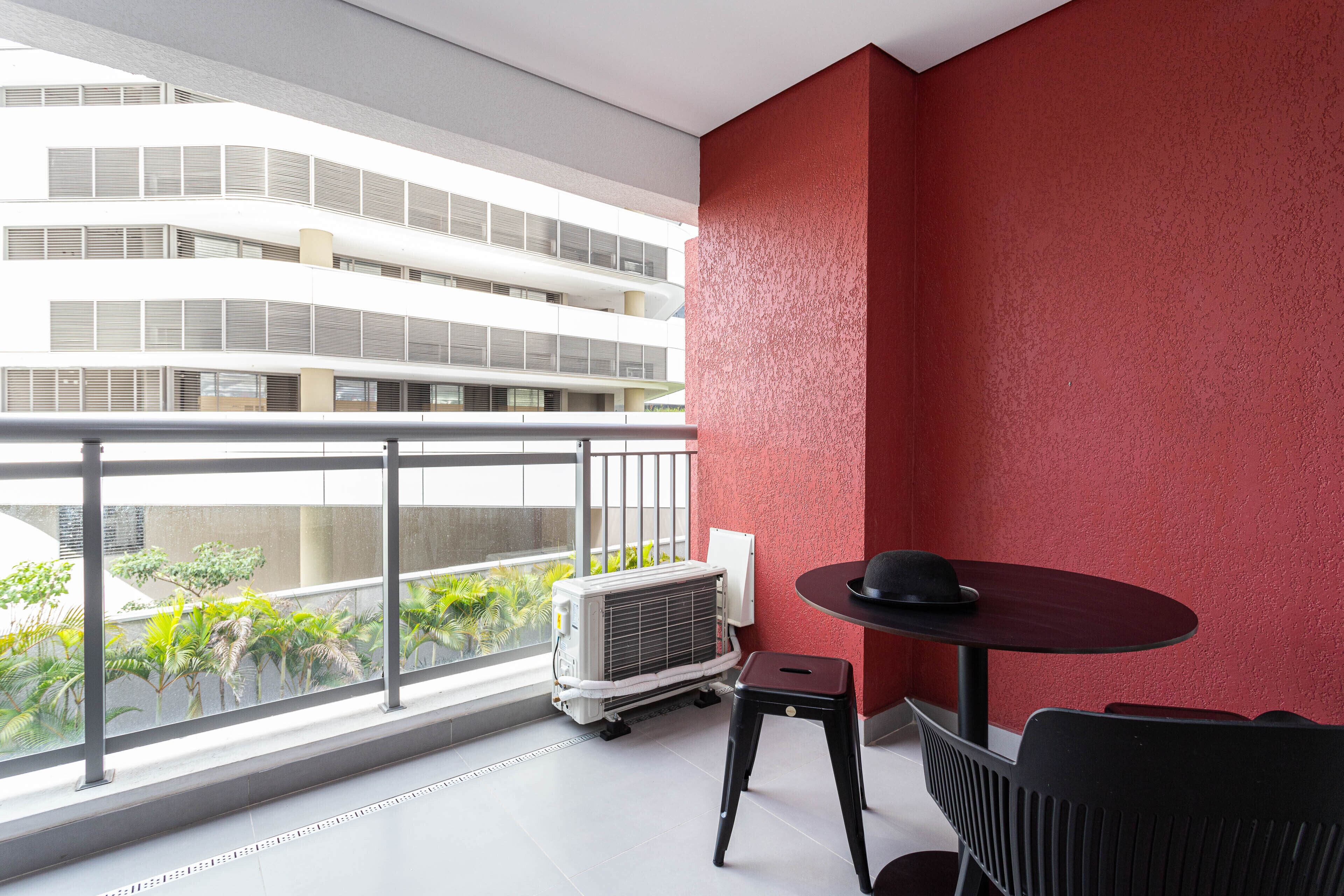 Un balcón con un muro rojo, una mesa con un sombrero y un equipo de aire acondicionado.
