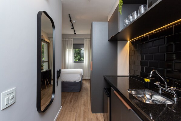 A compact hotel room with a kitchenette, a bed, a mirror, and a view of the outdoors.