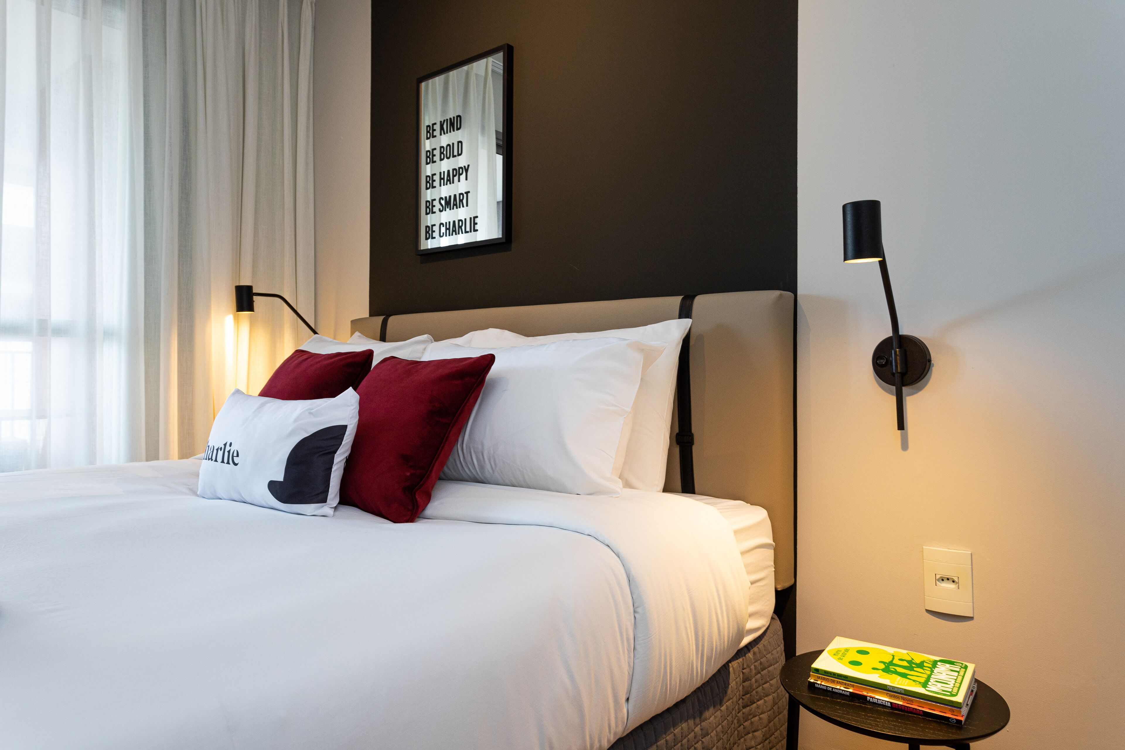A neatly made bed with white and red pillows, a motivational quote on the wall, a bedside table with books, and a lamp.