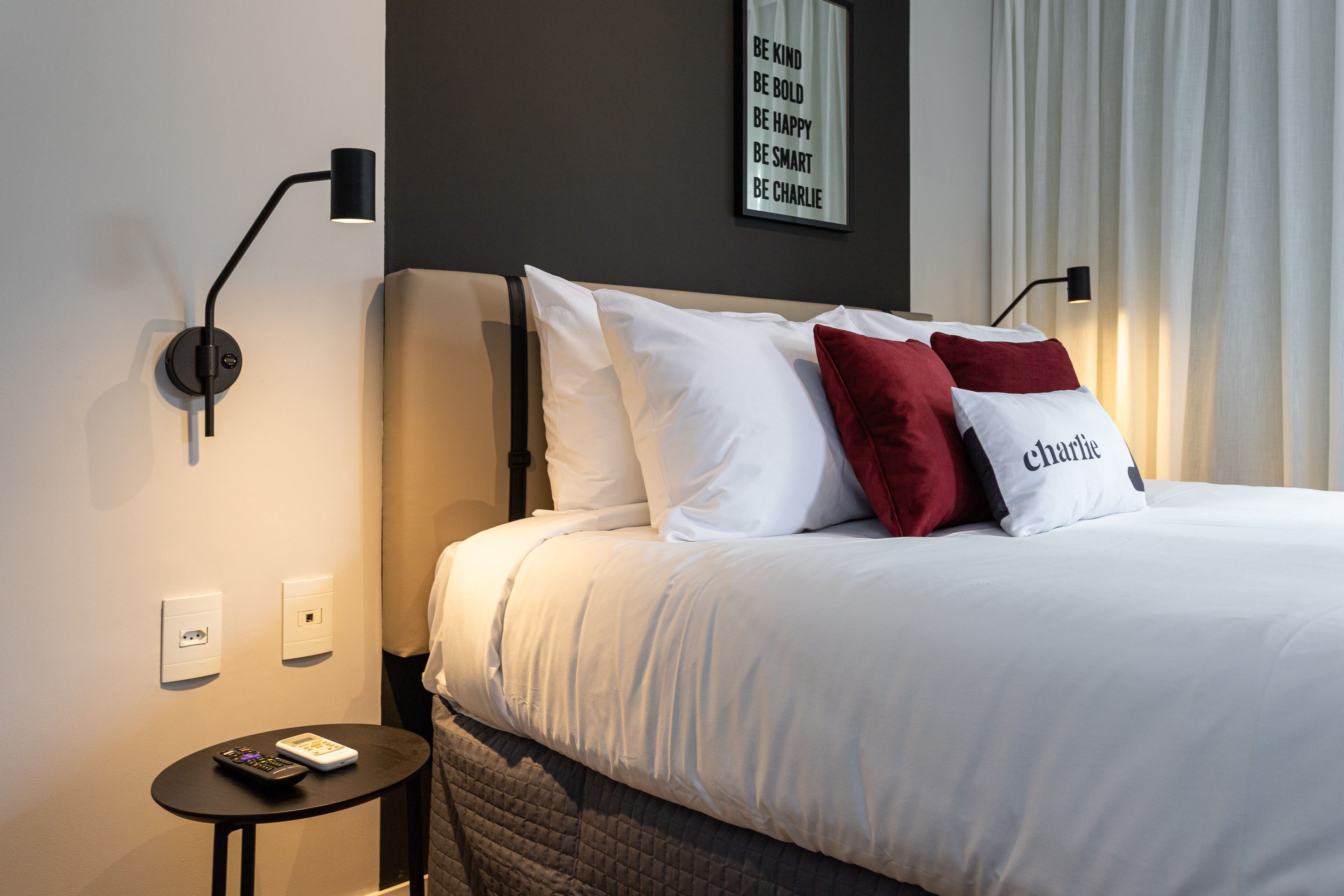 Ein ordentlich bezogenes Bett mit weißen und roten Kissen, ein kleiner Nachttisch mit Fernbedienung und ein motivierendes Zitat an der Wand.