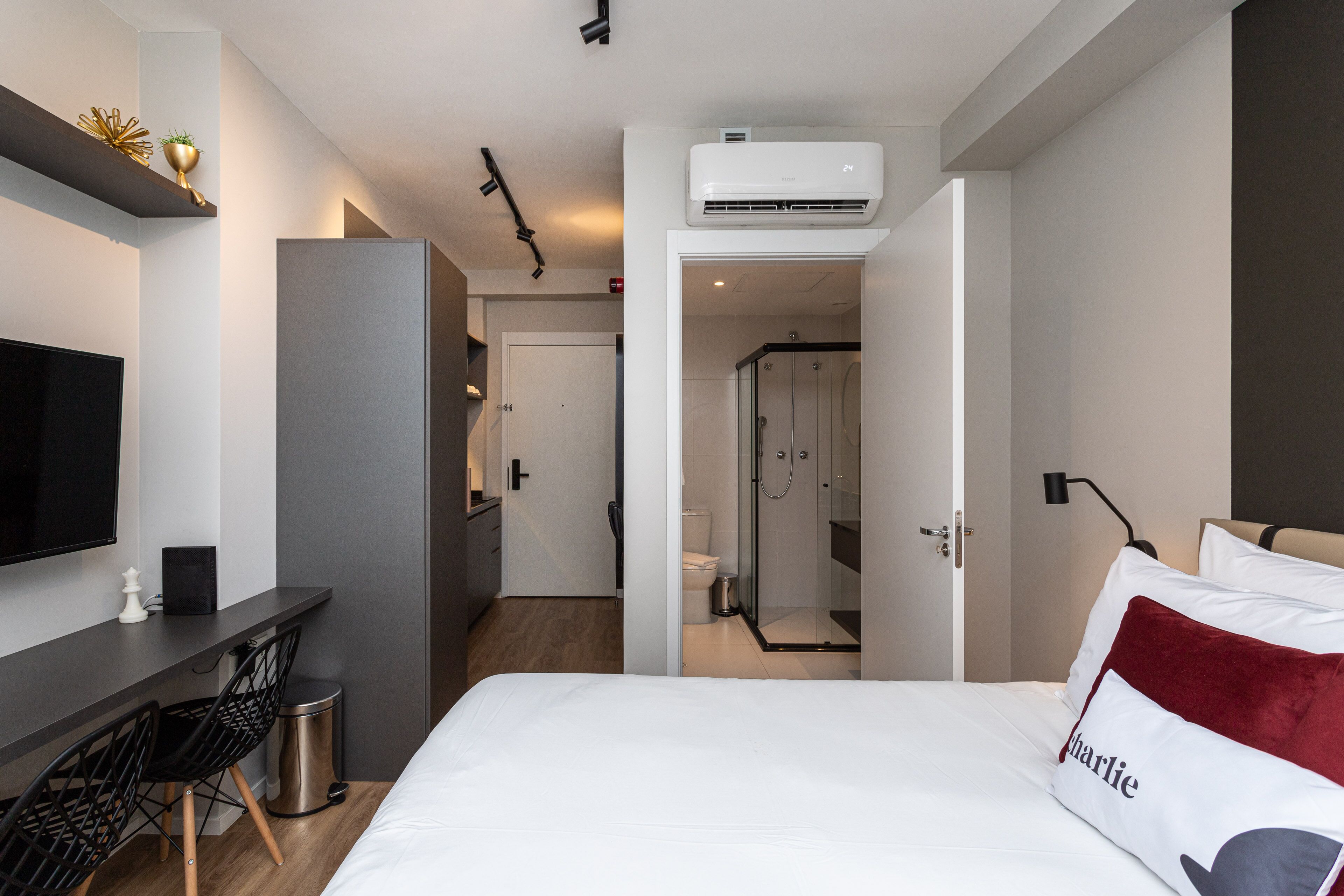 A modern hotel room with a bed, a desk with chairs, a TV, and an air conditioning unit.