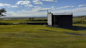 A small, single-story building with a sloped roof, situated in a grassy field with a clear sky and distant horizon. - Cosy Cabin Near Seljalandsfoss (Rangárþing eystra)