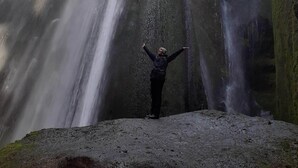 A person standing in front of a waterfall with arms raised, surrounded by rocky terrain and greenery. - Cosy Cabin Near Seljalandsfoss (Rangárþing eystra)