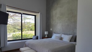 Quarto com uma janela grande, uma cama, um banco e uma mesa de cabeceira com abajur.