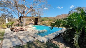 Quintal com piscina, cadeiras de descanso, pérgola e vista para colinas.