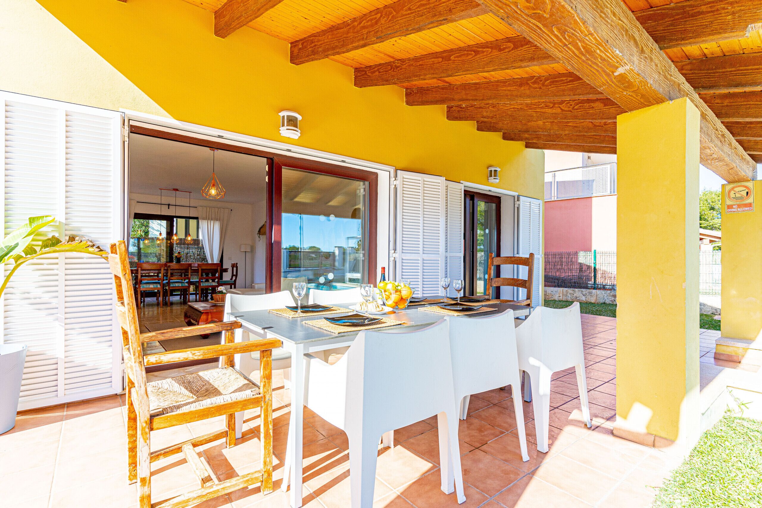An outdoor dining area with white tables and chairs, a wooden pergola, and a view of the interior through open doors.