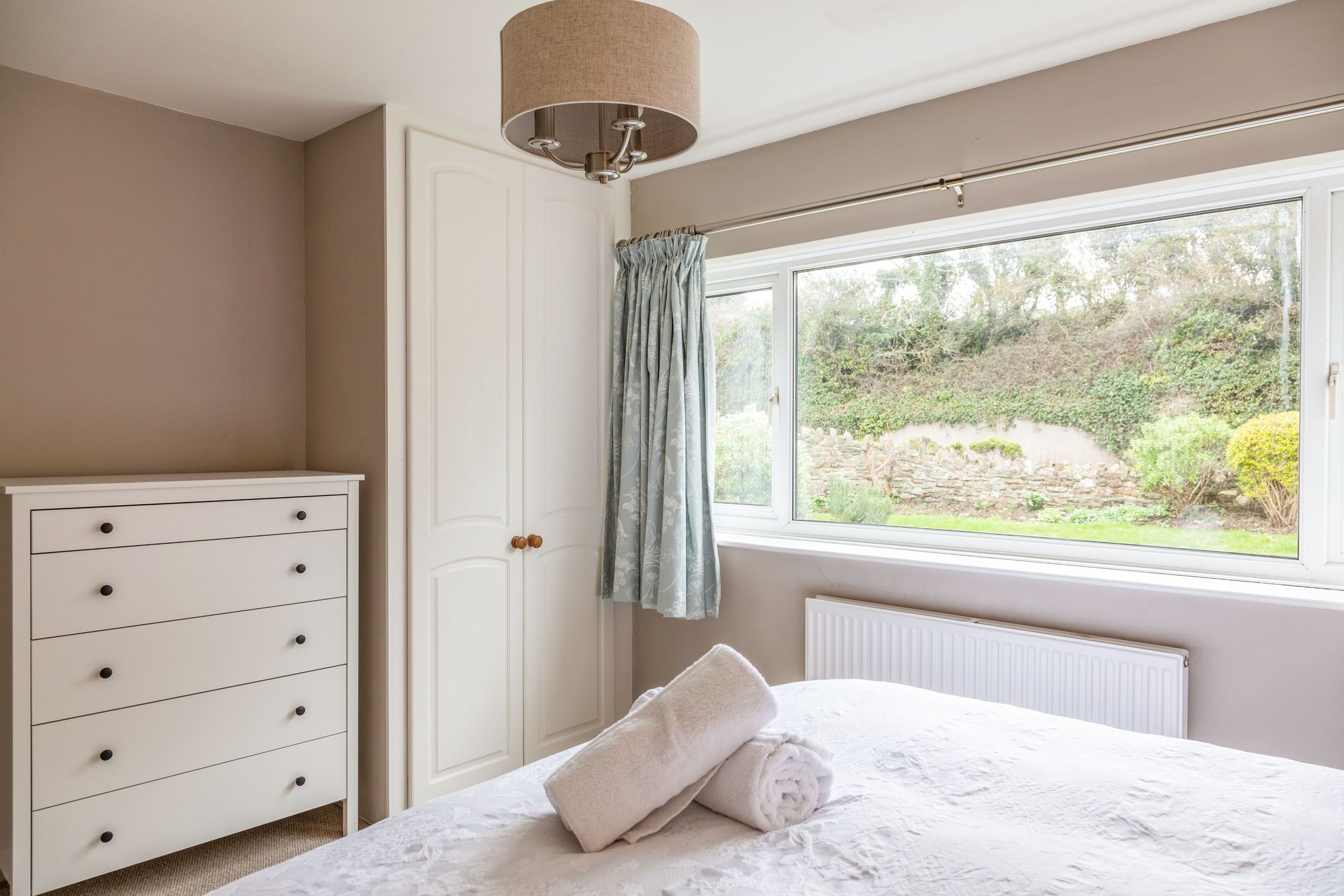A bedroom with a bed, a dresser, a window with a view of greenery, and a ceiling light fixture.