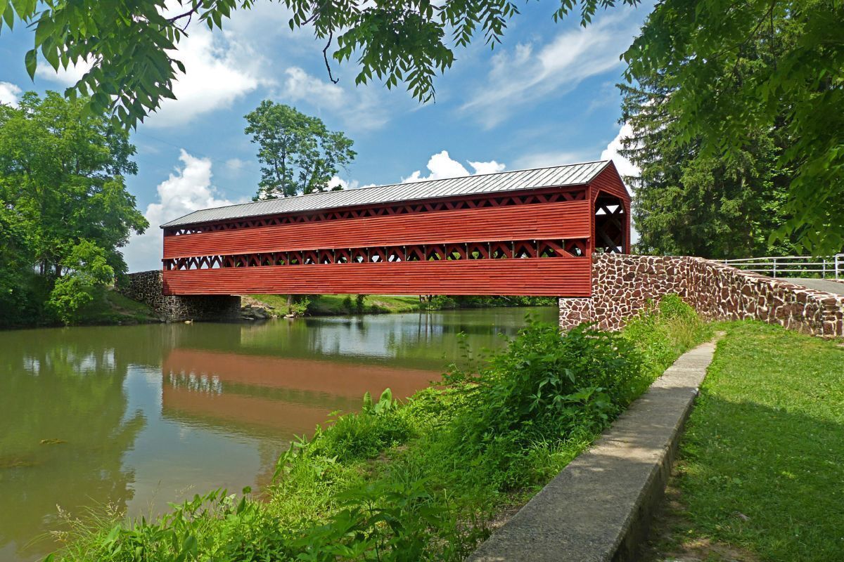 A red covered bridge over a calm river, surrounded by lush greenery and a clear blue sky.