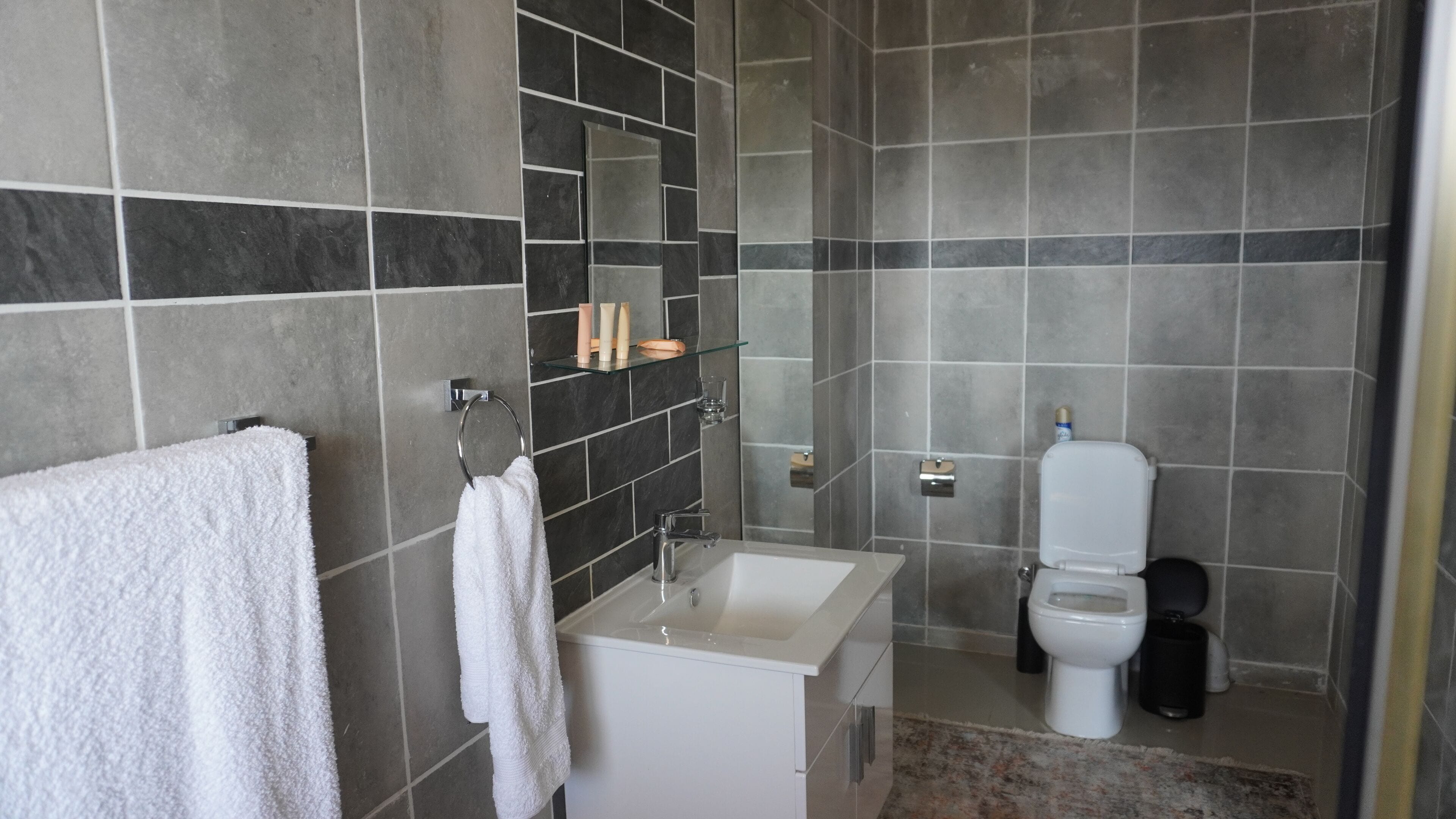 Uma casa de banho moderna com azulejos cinzentos, uma sanita branca, um lavatório e um compartimento de duche com divisória de vidro.