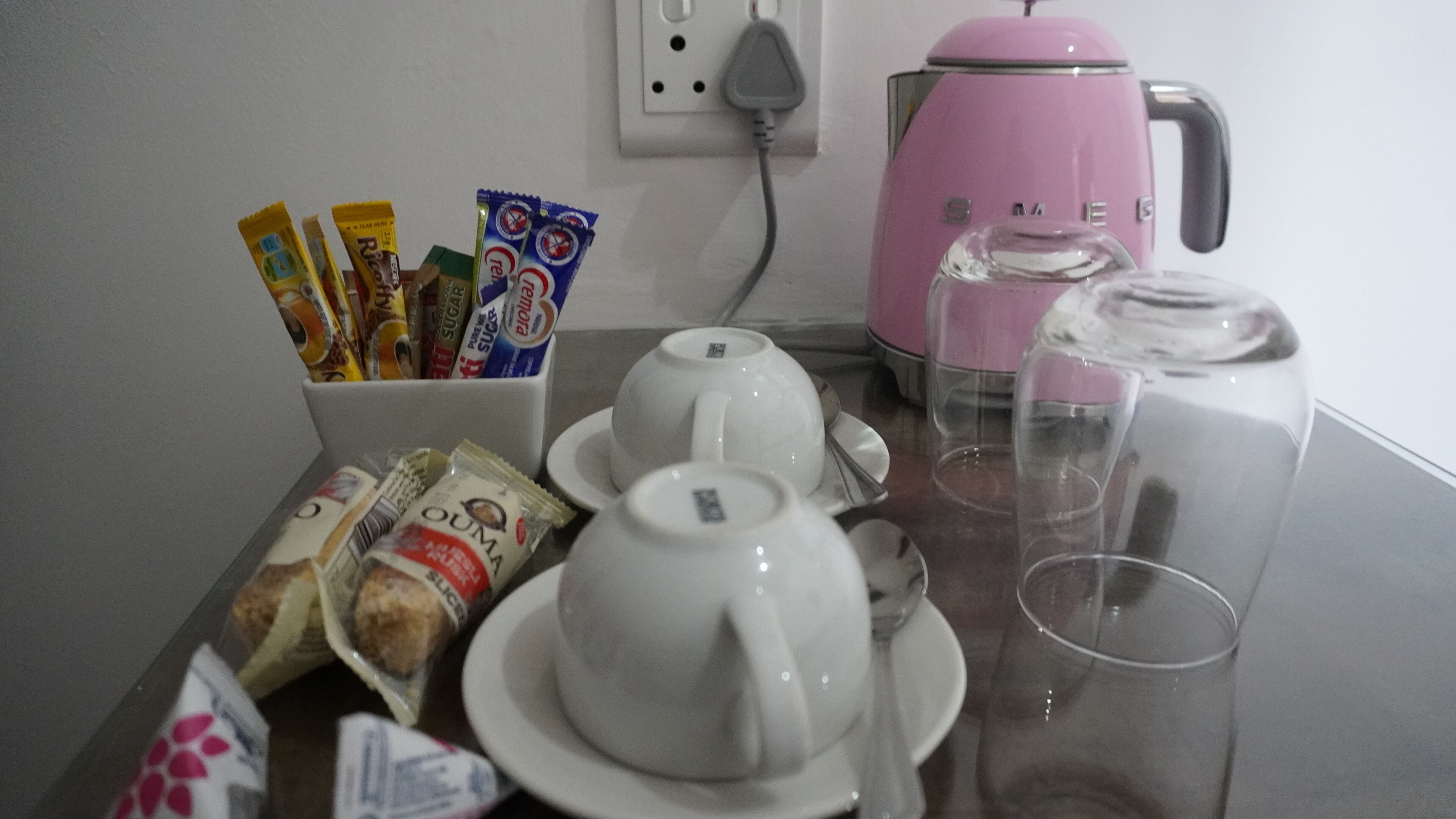 Une table avec une bouilloire rose, deux théières blanches, des tasses et un pichet en verre. On y trouve également des paquets de collations et un sachet de biscuits.