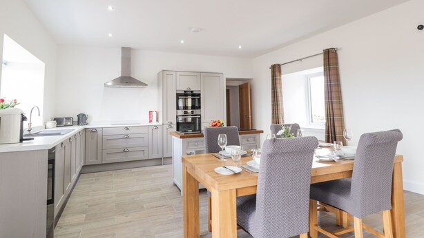 A modern kitchen with grey cabinets, a built-in oven, and a dining area with a wooden table and grey upholstered chairs. - Firthview (Nairn)