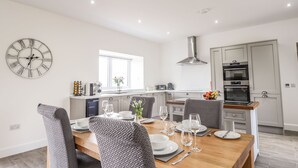 A modern kitchen with a built-in oven, a dining area with a wooden table and grey upholstered chairs, and a large wall clock. - Firthview (Nairn)
