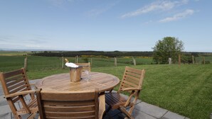 A round wooden table with chairs, a bucket, and a glass on it, set on a patio with a view of a grassy field and a tree. - Firthview (Nairn)