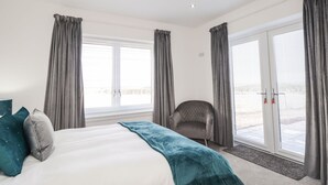 A modern bedroom with a large bed, grey curtains, a grey armchair, and a view through glass doors. - Firthview (Nairn)