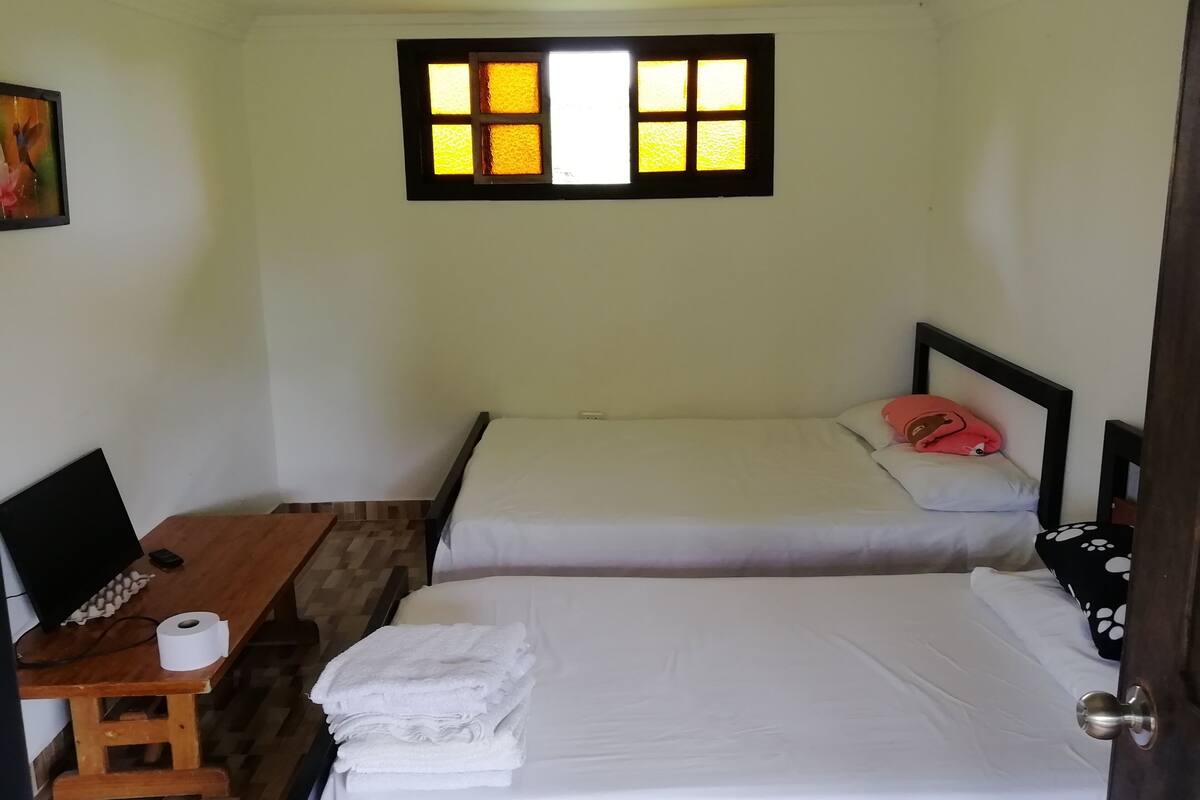 A small, clean room with two beds, a desk with a computer and a roll of toilet paper, and a window with a view of the outside.