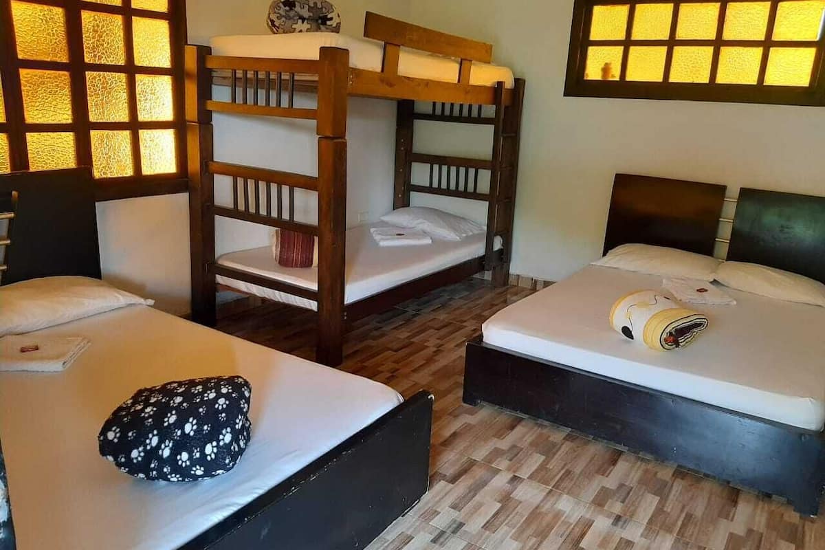 A room with two beds, one with a bunk bed and the other with a single bed. There is a wooden floor, a window with a view, and a wall with a decorative panel.