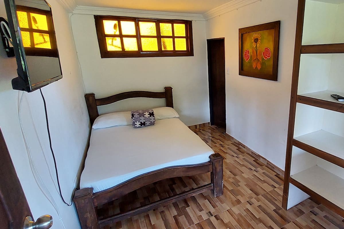 A bedroom with a wooden bed, a window with multiple panes, a painting on the wall, and a wooden shelf unit.
