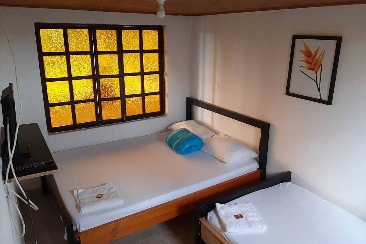 A single bed with a wooden frame, a small desk with a chair, a window with yellow-tinted glass, and a framed picture on the wall.