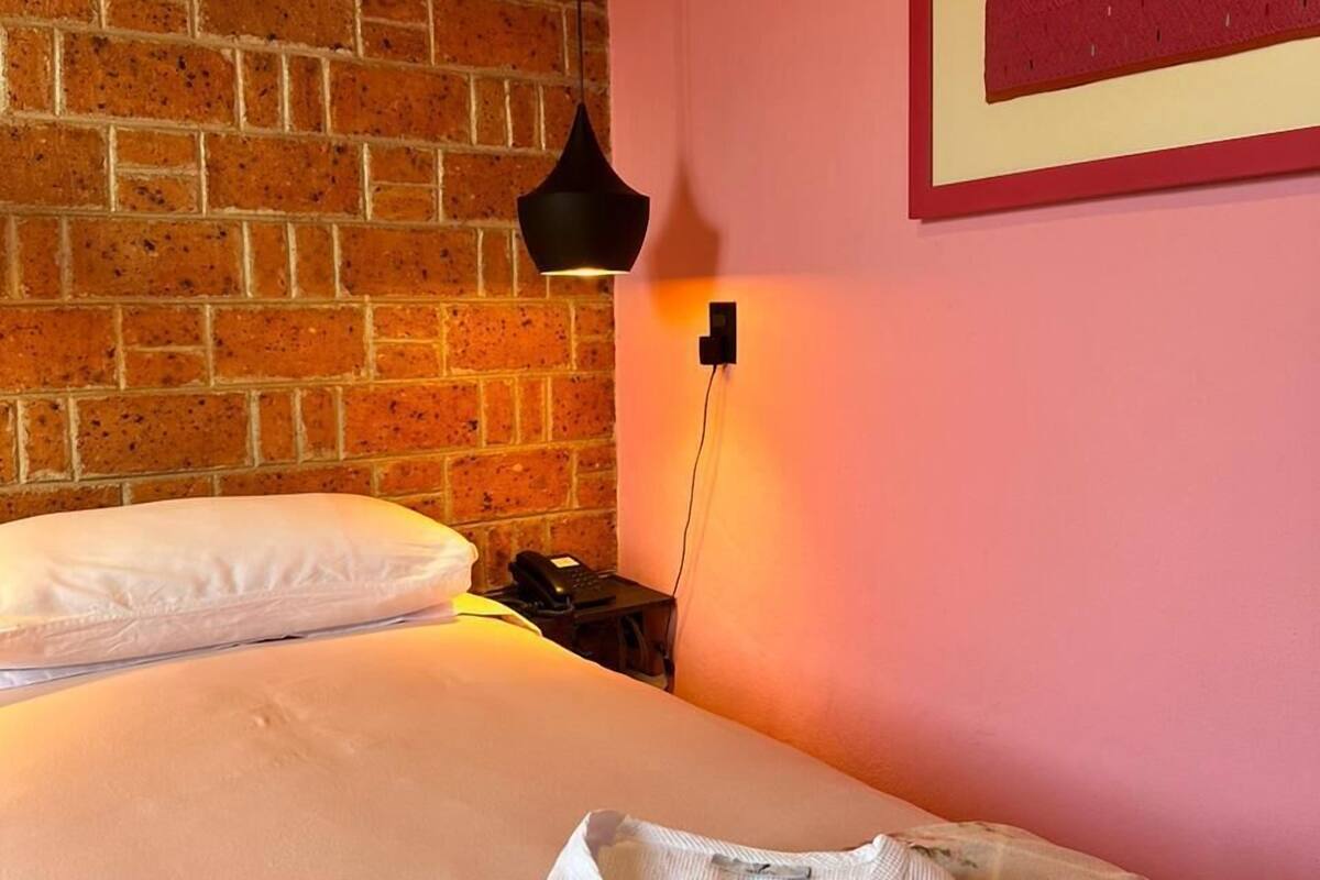 Ein Hotelzimmer mit Backsteinwand, einem Bett mit weißer Bettwäsche, einem Nachttisch mit Lampe und einem gerahmten Bild an der Wand.