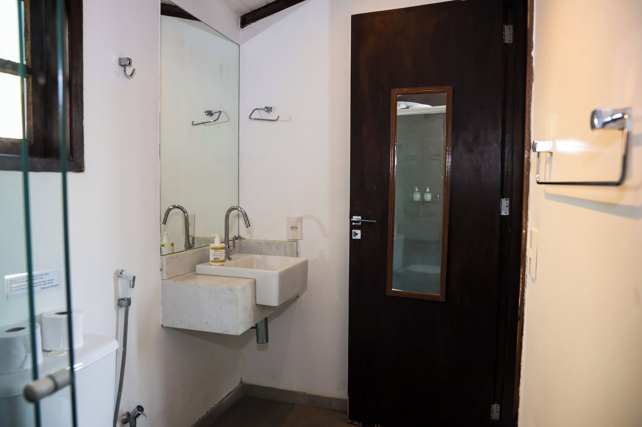 Un baño con un lavamanos blanco, dos espejos, una puerta de madera oscura y una mampara de cristal.