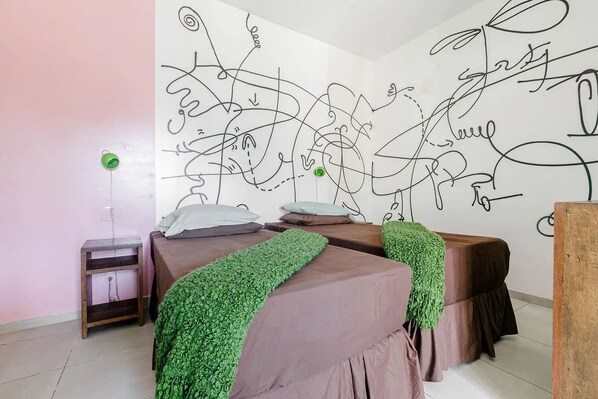 A bedroom with two beds, a wall with abstract line art, a bedside table with a lamp, and a small wooden nightstand.