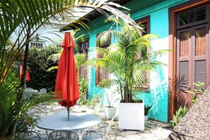 Un patio con mesa y sillas, rodeado de plantas en macetas y un paraguas rojo.