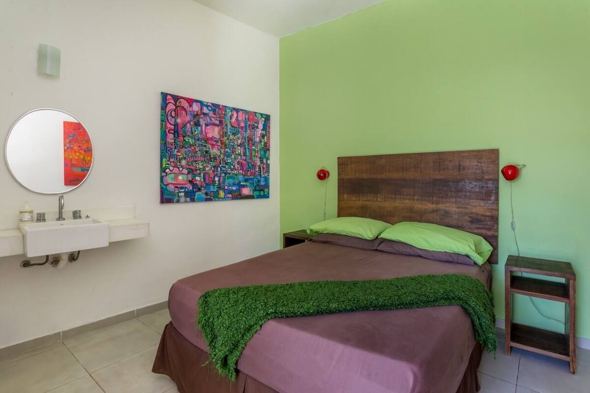 A bedroom with a bed, a nightstand, a bedside lamp, a mirror, and a colorful abstract painting on the wall.
