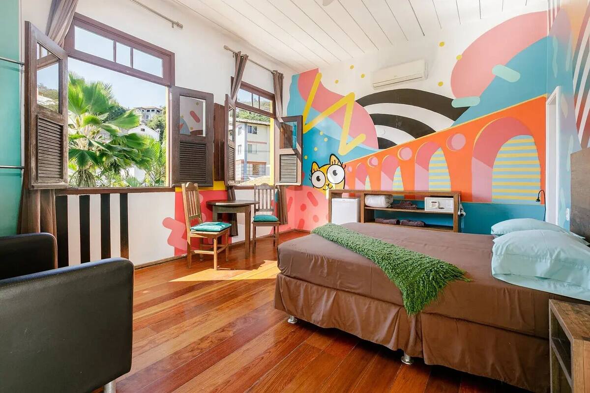 A bedroom with a bed, a desk, and a chair. There are windows and a colorful wall mural.