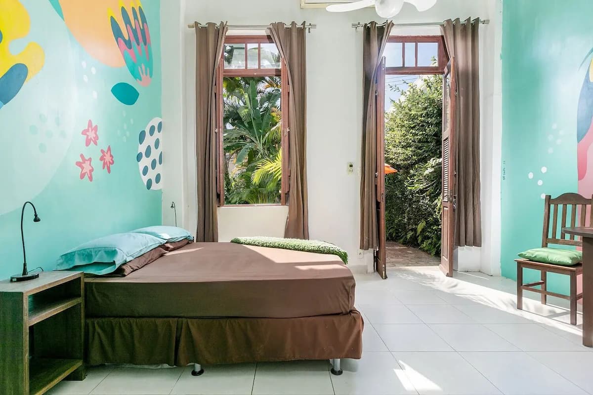 A room with a bed, a desk, and a chair. There are two windows with curtains, and a view of greenery outside.