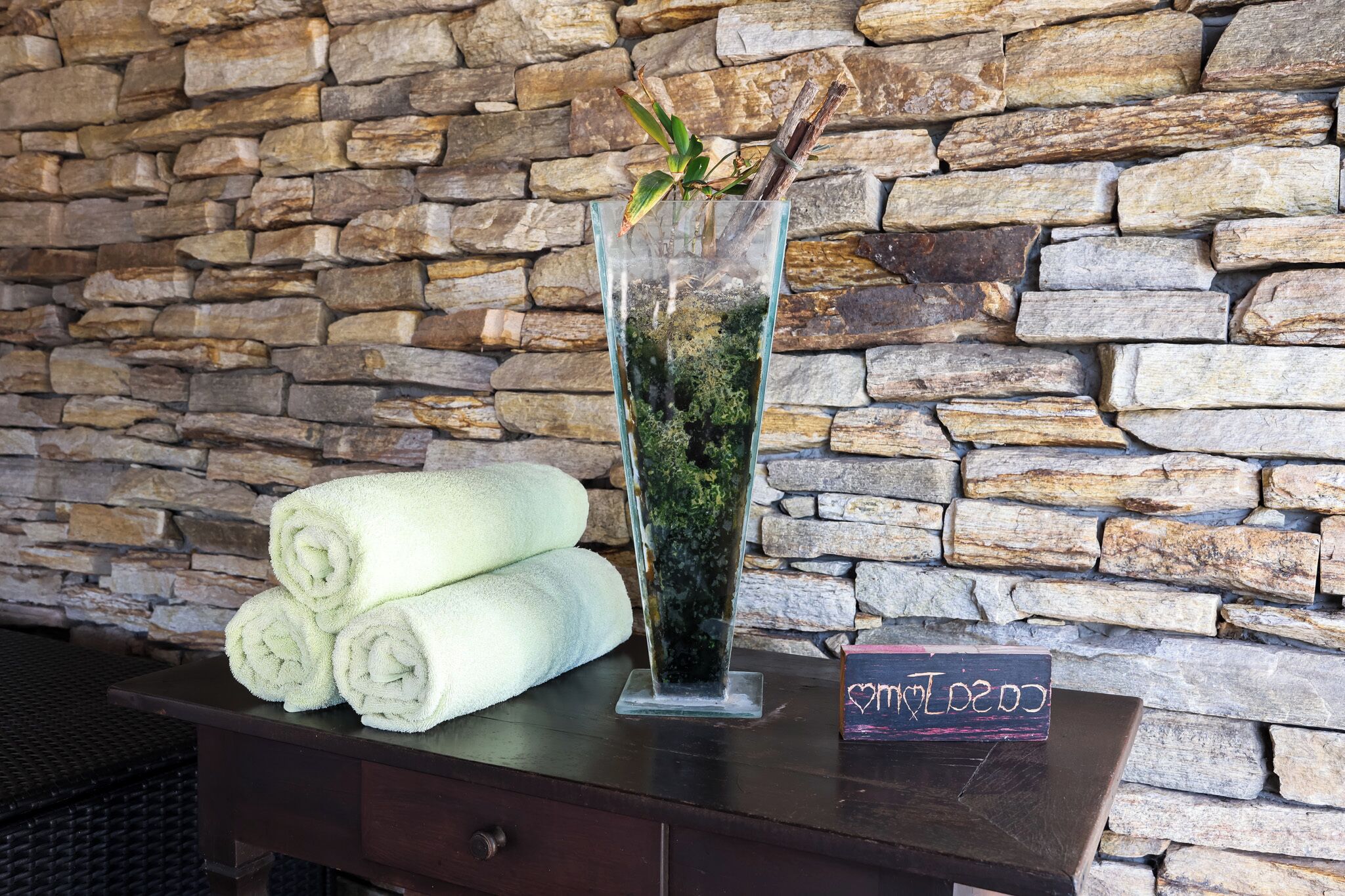 A glass vase with moss and twigs, a stack of white towels, and a sign with a heart symbol on a wooden table against a stone wall.