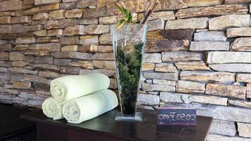 A glass vase with moss and twigs, a stack of white towels, and a sign with a heart symbol on a wooden table against a stone wall.