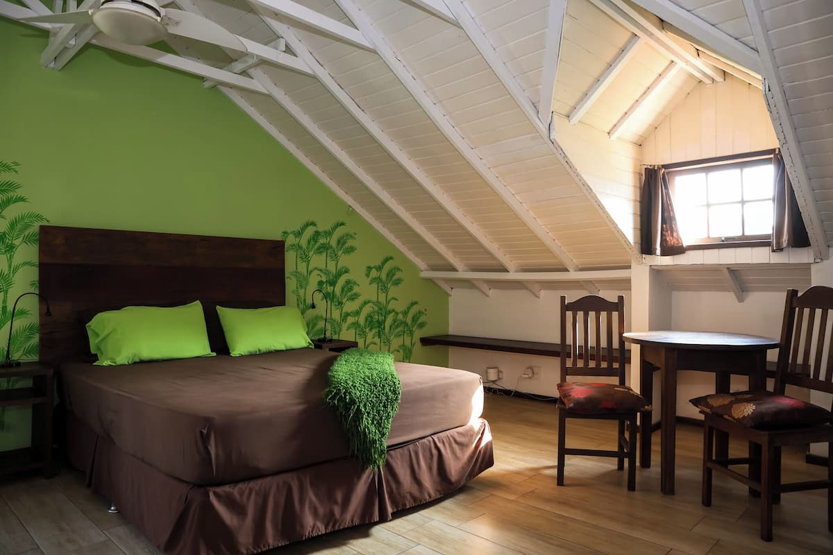 A bedroom with a bed, green walls, a wooden headboard, a ceiling fan, a window with curtains, and a dining area with chairs and a table.