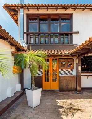 A building with a tiled roof, a wooden door, and a small outdoor seating area with a white planter and palm tree.