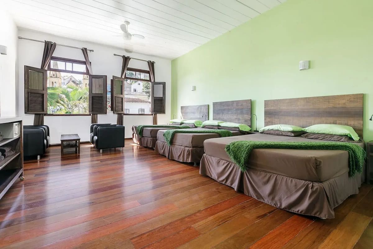 A hotel room with two beds, a wooden floor, a window with shutters, and a ceiling fan.