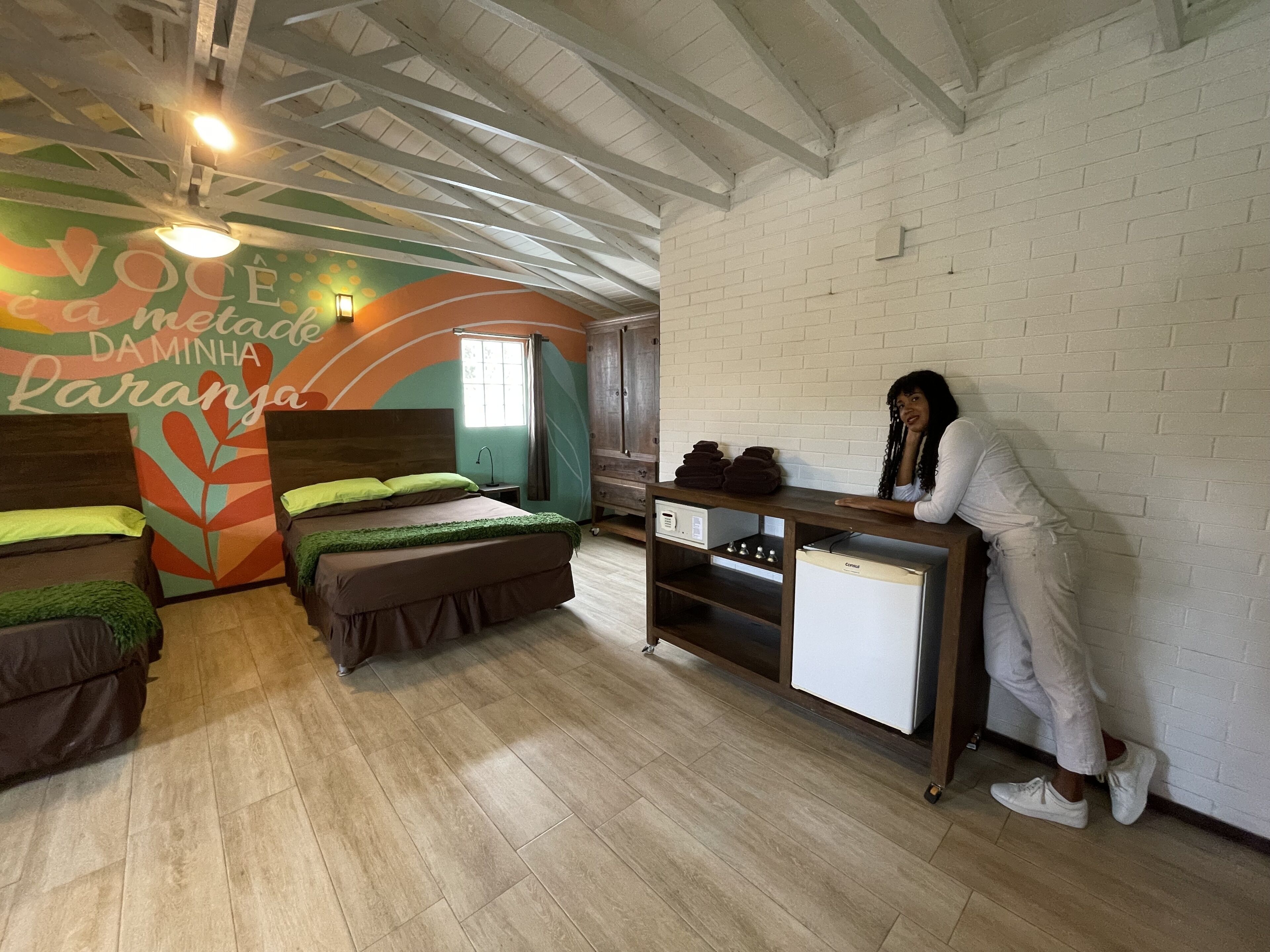 Une chambre avec deux lits, une petite table, un réfrigérateur et un mur orné d’une fresque colorée.