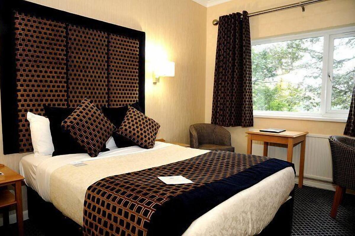 A hotel room with a large bed, a headboard with a geometric pattern, a small table, a chair, and a window with curtains.