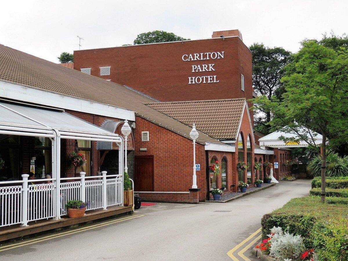 Hotel Carlton Park de ladrillo rojo con fachada blanca, zona de estar exterior cubierta y un camino de jardín.