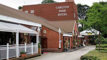 A red brick Carlton Park Hotel with a white facade, a covered outdoor seating area, and a garden pathway.