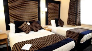 Habitación de hotel con dos camas, cada una con una colcha y almohadas estampadas, una mesita de noche y un ventanal con cortinas.