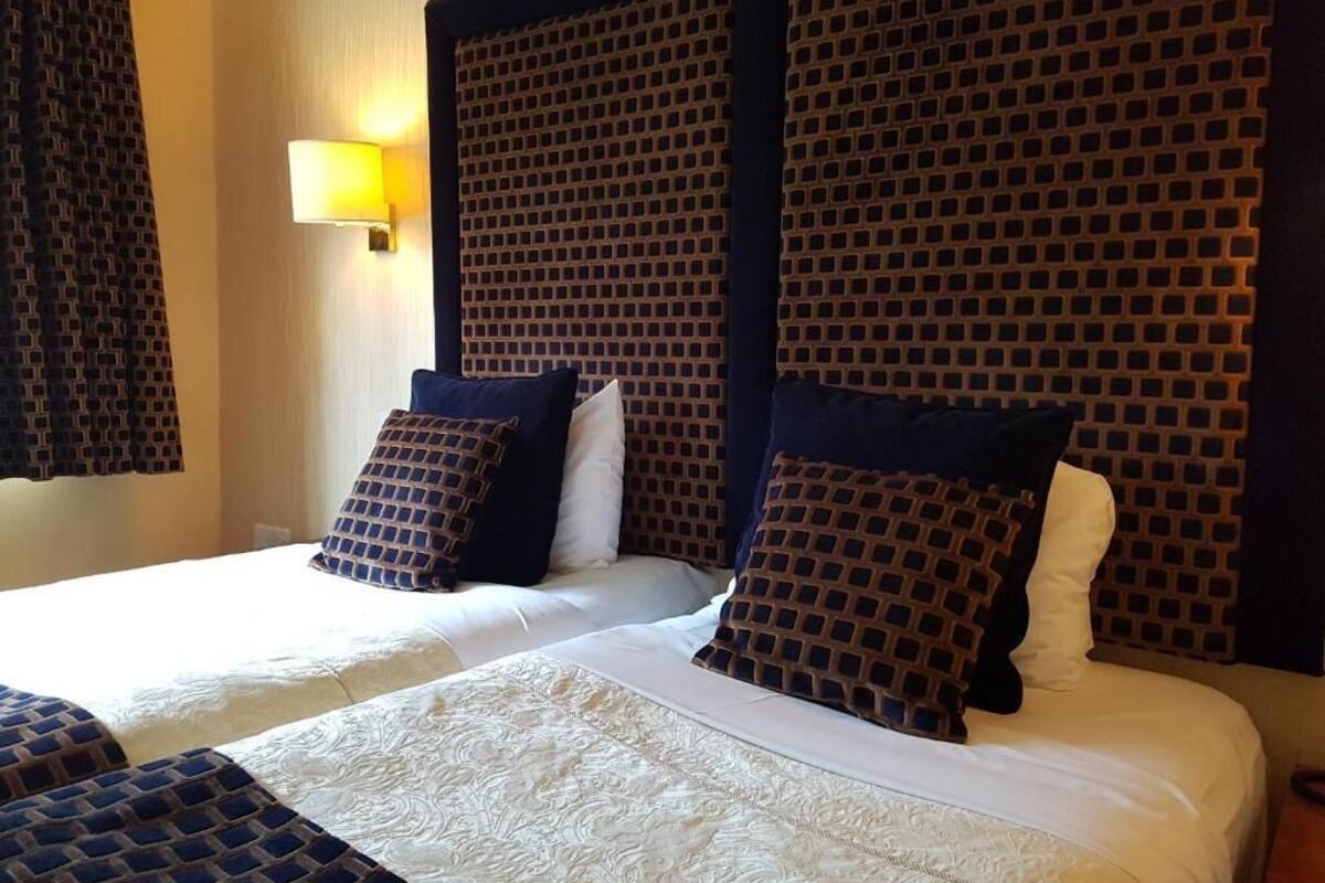 A hotel room with two beds, a patterned headboard, a wall-mounted lamp, and a patterned curtain.