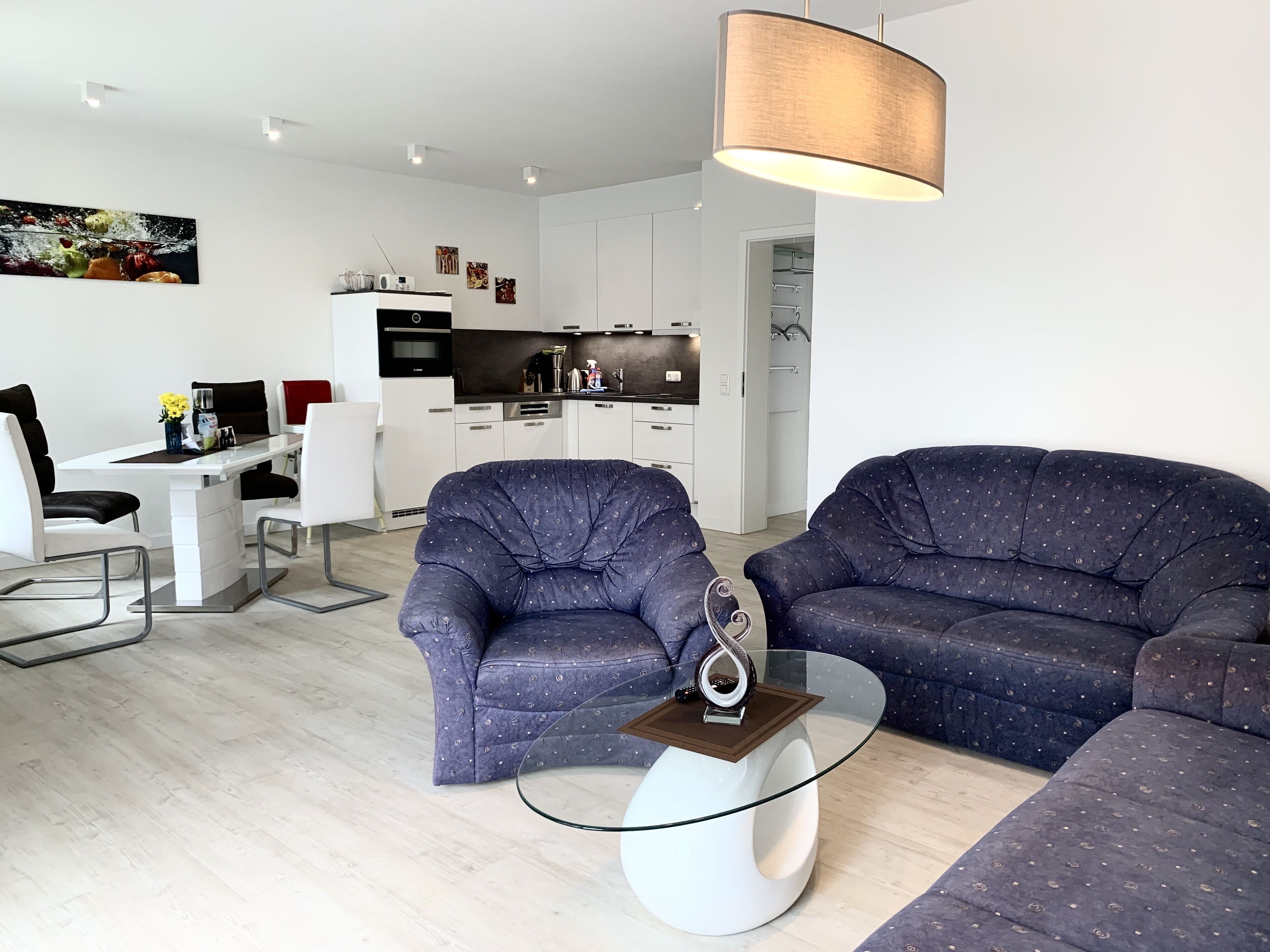 A modern living room with a glass coffee table, two patterned armchairs, a dining area with a white table and chairs, and a kitchen in the background.