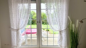 Ein Zimmer mit einer Schiebetür aus Glas, weißen Vorhängen, einem Topfpflanzen und Blick auf einen Außenbereich mit einem roten Tisch und Stühlen.