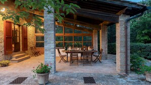 Une terrasse extérieure avec un pilier en briques, une table et des chaises en bois, le tout entouré de grandes fenêtres et de verdure.
