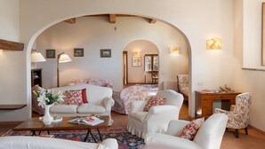 Un salon chaleureux avec un canapé, des fauteuils et une table basse. On y trouve un bureau avec une chaise et une lampe. La pièce est dotée d’un plafond en arc de cercle et les murs sont ornés de tableaux encadrés.