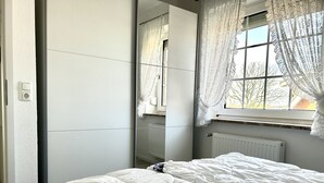 A bedroom with a bed, a window with lace curtains, a radiator, and a mirror reflecting the room. - Apartment with a view of the dyke for up to 4 people. -Turquoise (Büsum)