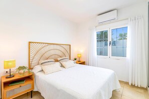 Un dormitorio con cama, mesita de noche con lámpara, una planta pequeña y ventana con cortinas.