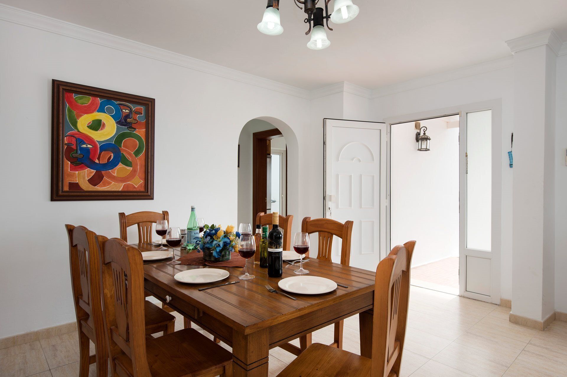 A dining area with a wooden table set for four, surrounded by chairs. There is a colorful abstract painting on the wall, a chandelier above the table, and a door leading to another room.