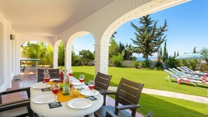 An outdoor patio with a table set for dining, surrounded by chairs, and overlooking a well-maintained lawn and a view of the ocean. - Villa Mi Sueno by Villa Plus (La Capellania)