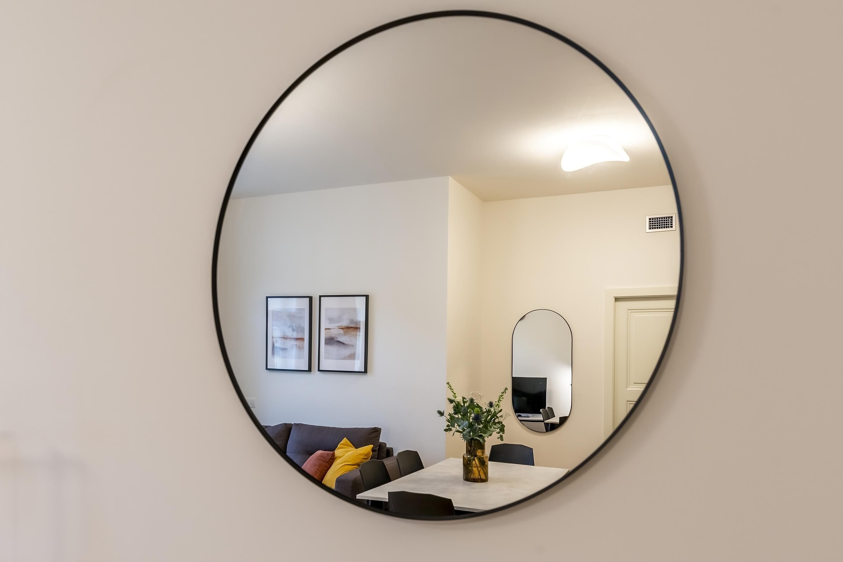 A round mirror reflecting a living room with a sofa, a coffee table with a vase, and two framed pictures on the wall.